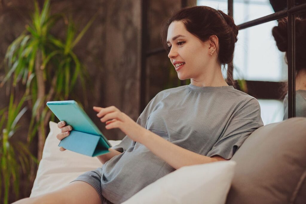 young woman seated comfortably and reviewing the 5 benefits of telehealth on her wireless tablet