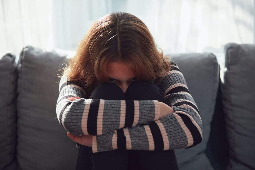 troubled young woman sitting on couch with knees pulled up to chest and arms wrapped around herself attempting to understand what is high functioning anxiety