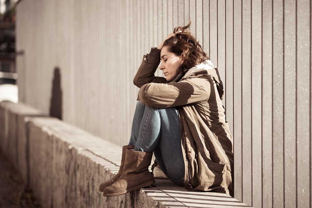 weary and distressed woman sitting outside on concrete wall leaning against industrial fence displaying common signs of trauma in adults