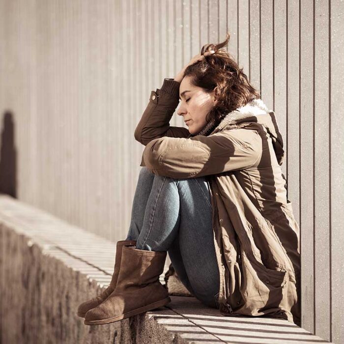 weary and distressed woman sitting outside on concrete wall leaning against industrial fence displaying common signs of trauma in adults