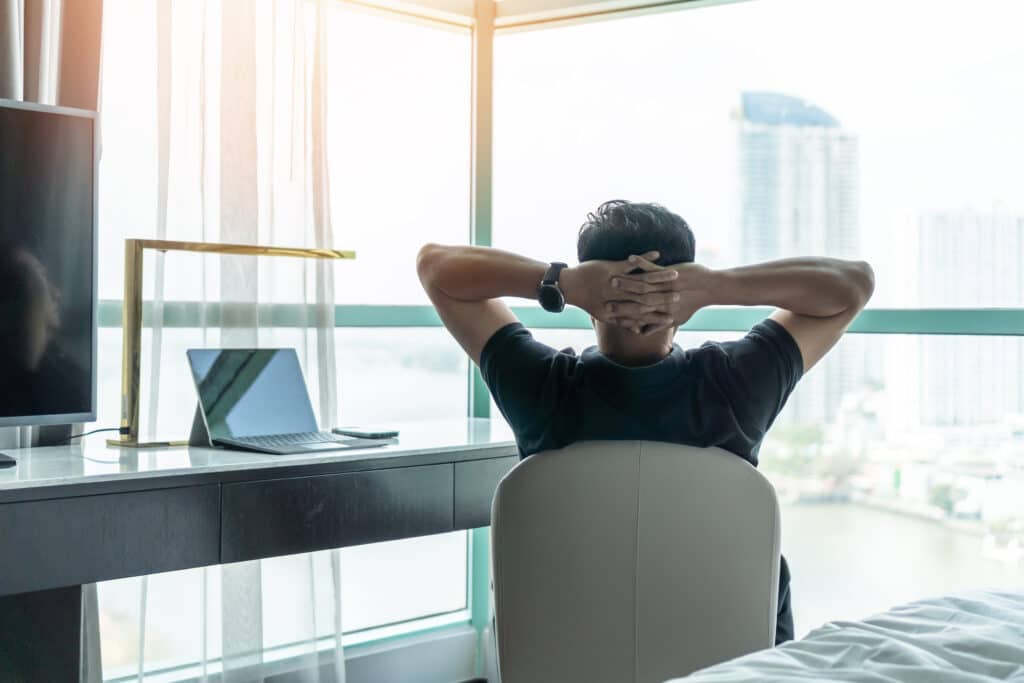 man sitting at desk with arms crossed behind his head staring longingly out his high rise window who is struggling with work-life balance