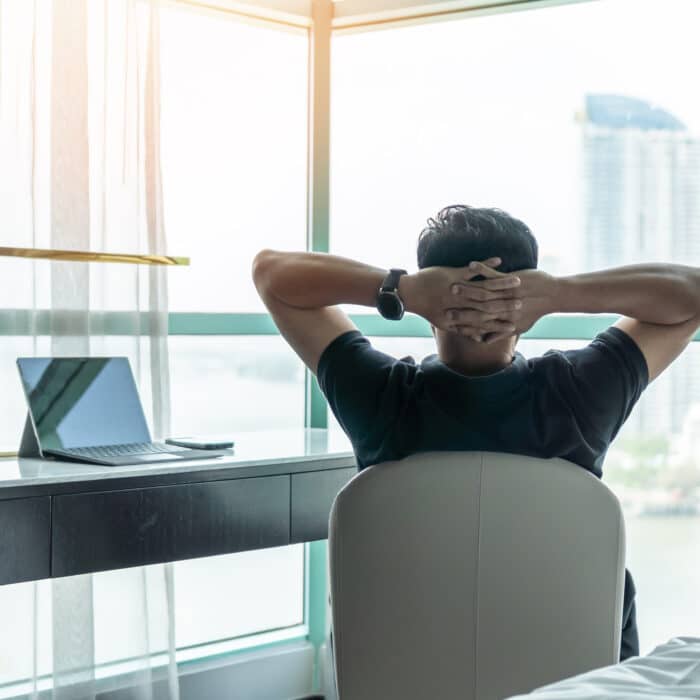 man sitting at desk with arms crossed behind his head staring longingly out his high rise window who is struggling with work-life balance